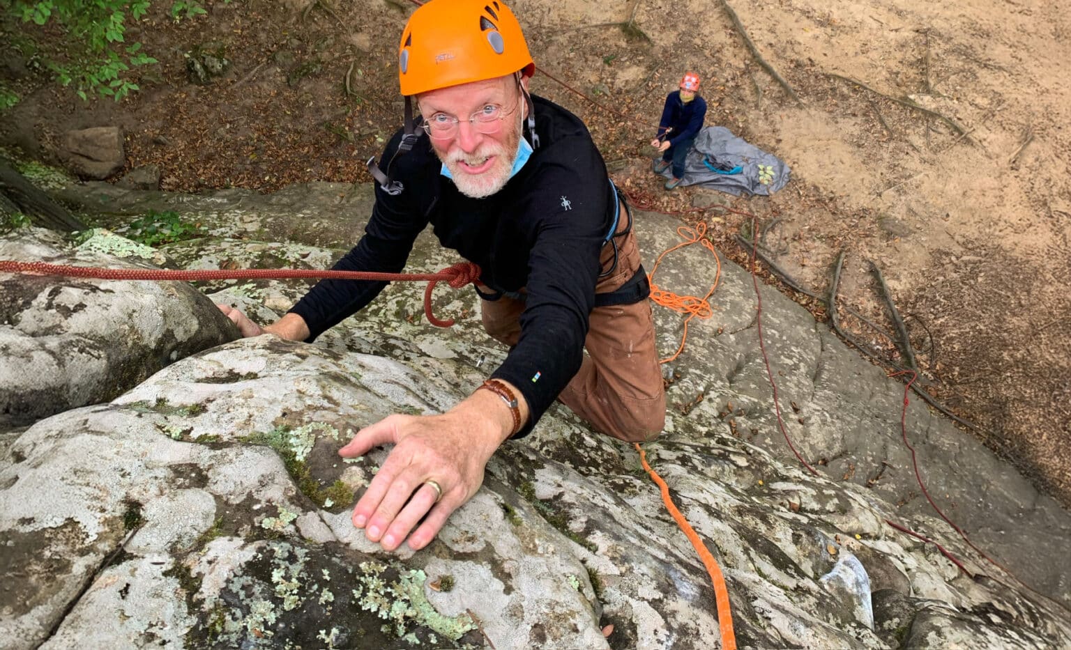 Older climber wearing an orange helmet and glasses ascending a rocky cliff face, gripping textured stone while secured by an orange rope, with a belayer standing below on a dirt and leaf-covered slope.