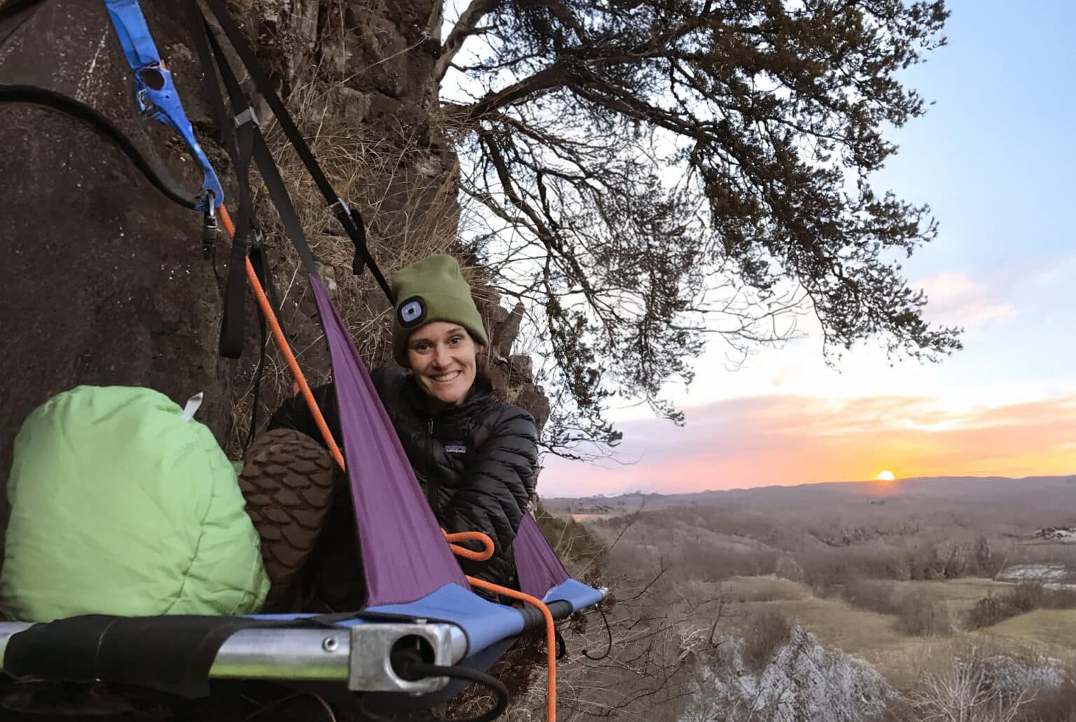 Person bundled in outdoor gear smiling while sitting on a portaledge attached to a cliff, with climbing ropes secured above and a colorful sunset glowing over rolling hills in the distance.