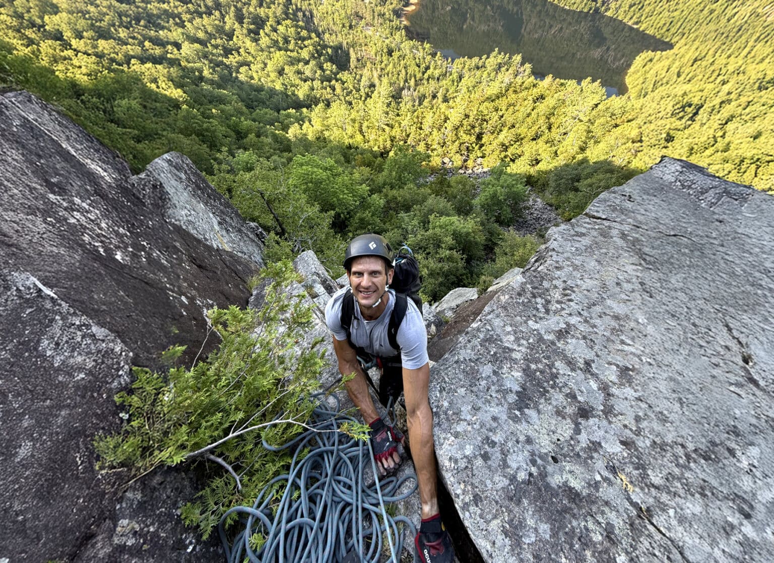 Climber wearing a helmet and gloves smiling while topping out on a rocky ledge, surrounded by thick green forest and overlooking a distant lake far below.