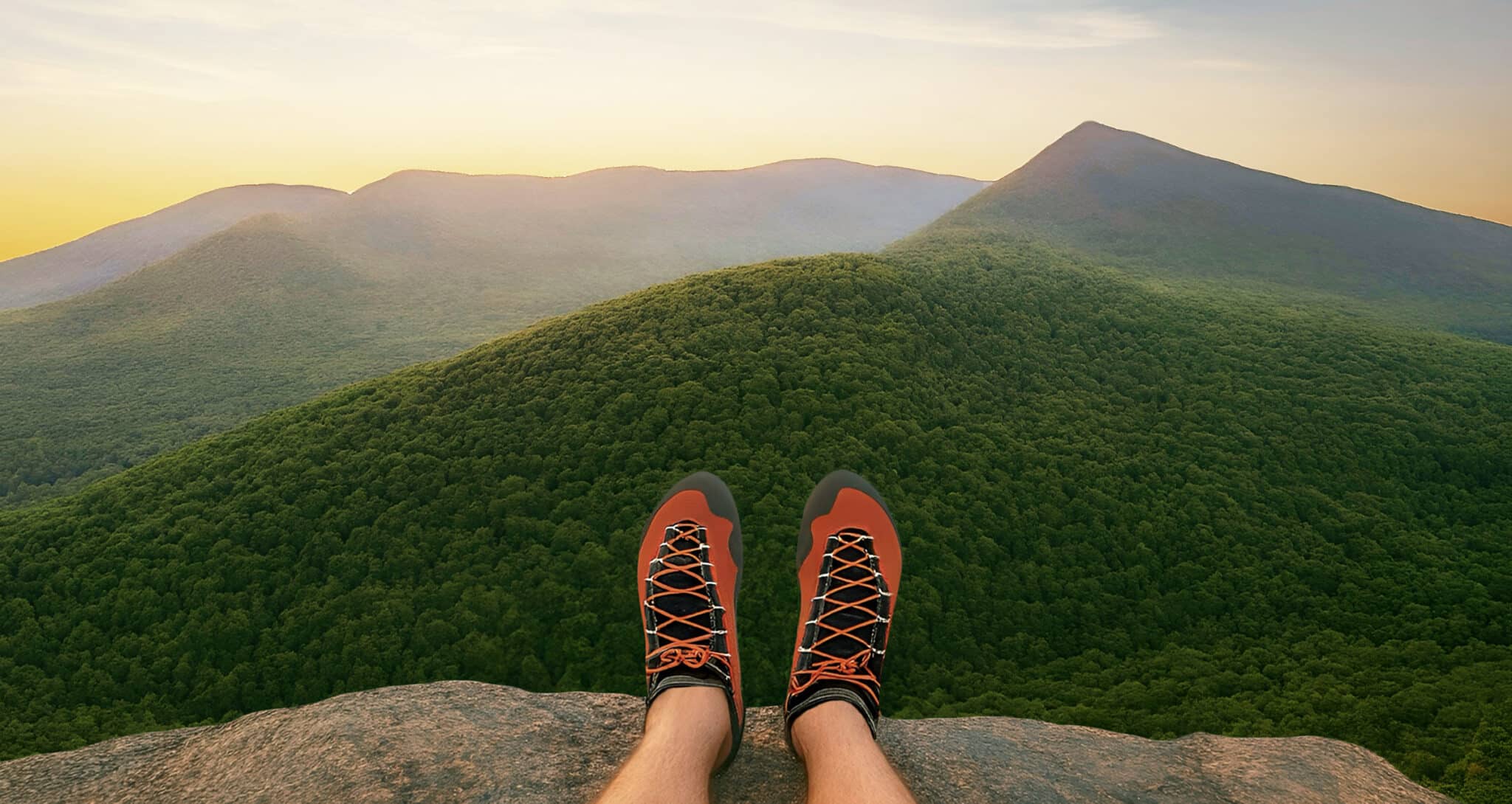 Person sitting on a rocky ledge wearing red climbing shoes, overlooking a lush green mountain range at sunset.