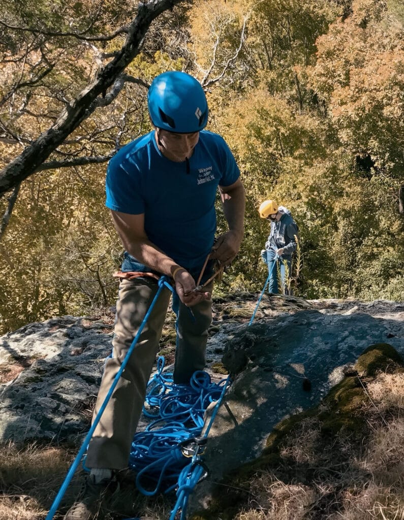 A man in a blue helmet and shirt setting up ropes and anchors on a rocky ledge with another climber preparing in the background.
