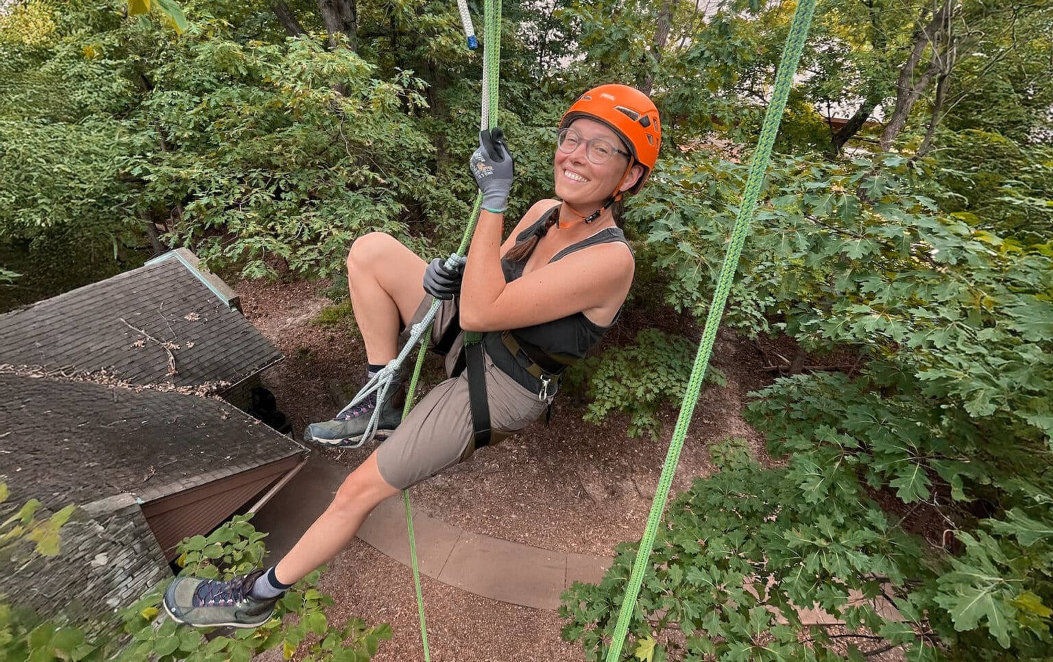 Person wearing an orange climbing helmet, harness, and gloves smiling while suspended on a rope high in a tree, with leafy branches and a rooftop visible below.