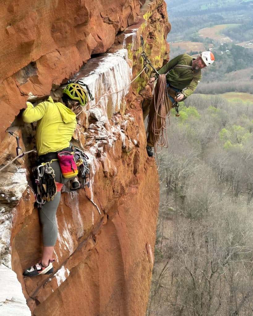 Two climbers on a steep red rock face, one actively climbing while the other manages rope and gear from a secure anchor position.