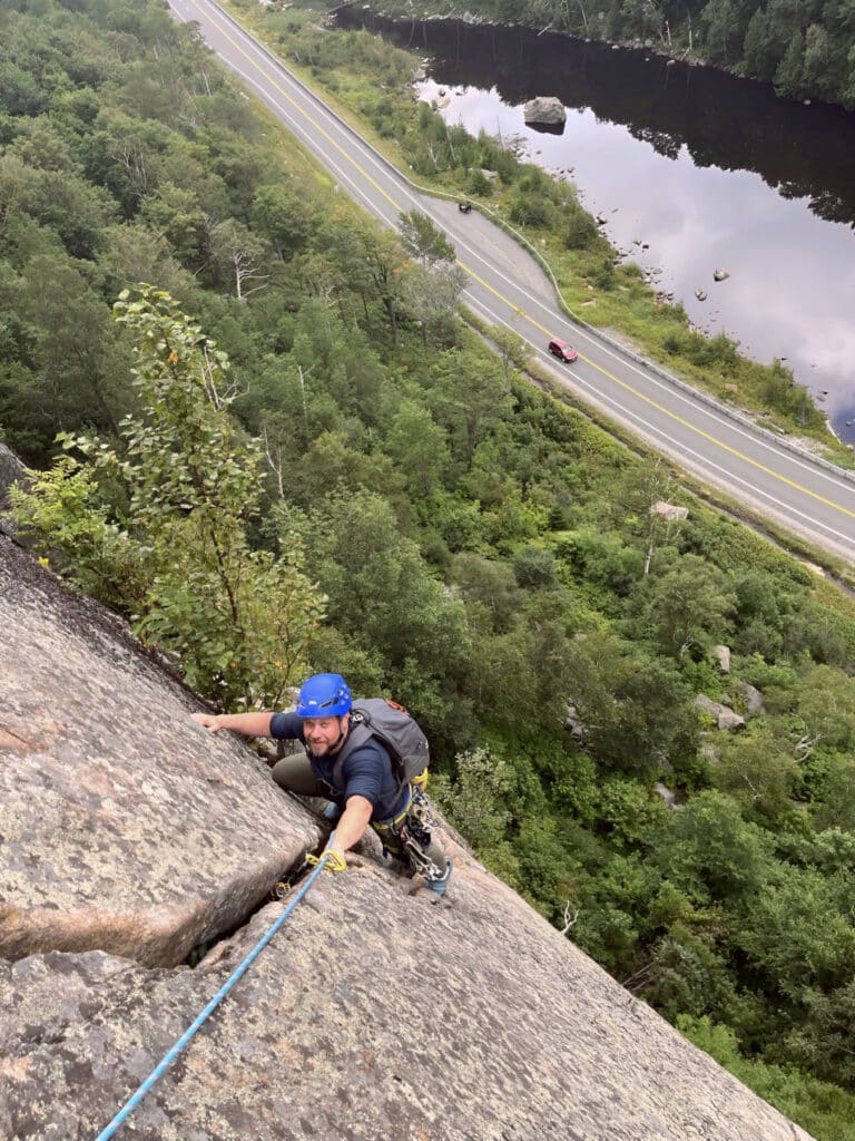 Climber wearing a blue helmet and backpack ascends a steep rock face with rope protection, overlooking a forest, highway, and river below.