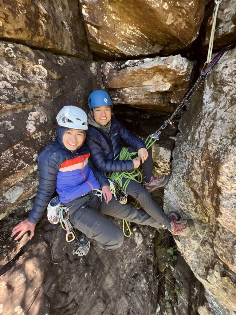 Two climbers wearing helmets and harnesses smile while sitting at a belay station on a rocky ledge, secured with ropes and gear.