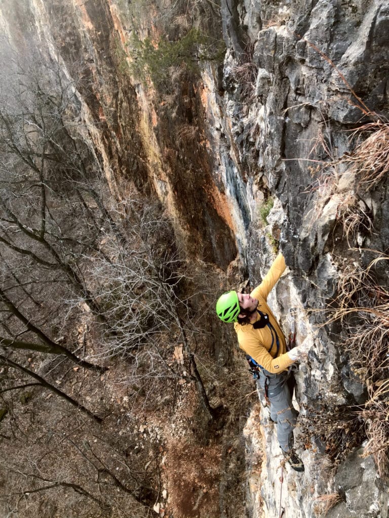 Climber wearing a green helmet and yellow jacket ascends a steep rocky cliff, surrounded by bare trees and rugged terrain below.