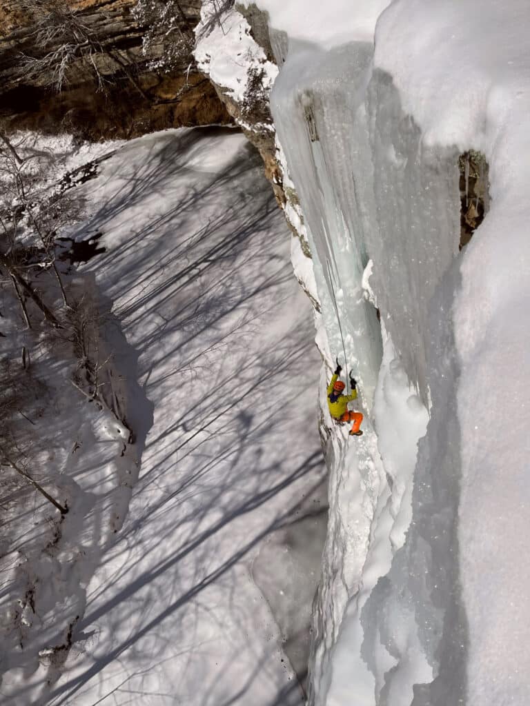 Ice climber wearing a yellow jacket and orange pants ascends a frozen waterfall with ice axes, surrounded by snow-covered cliffs and trees.