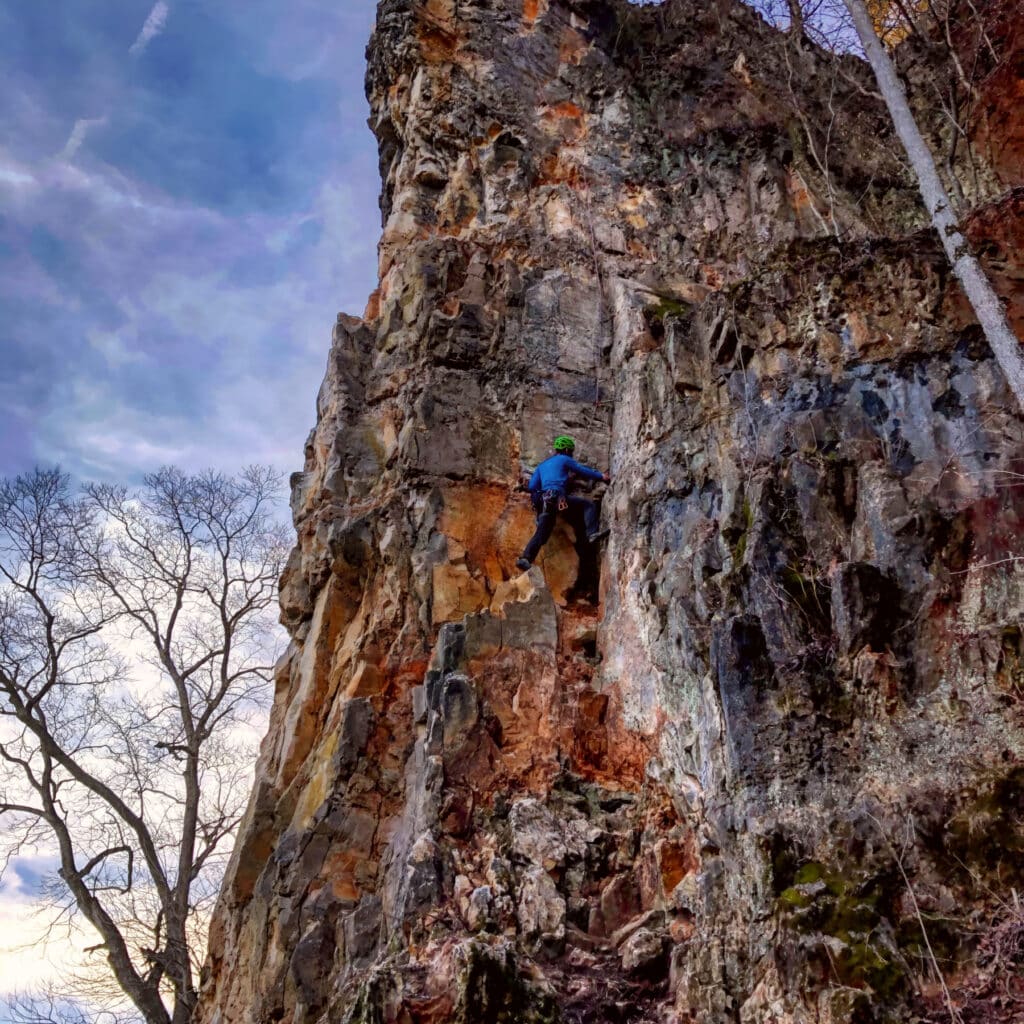 Climber wearing a green helmet and blue jacket scales a tall, rugged rock face against a backdrop of bare trees and a cloudy sky.