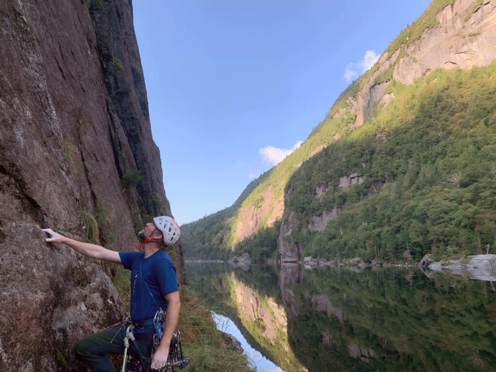 Climber wearing a white helmet and blue shirt prepares to ascend a tall rock wall beside a calm reflective lake surrounded by forested cliffs.