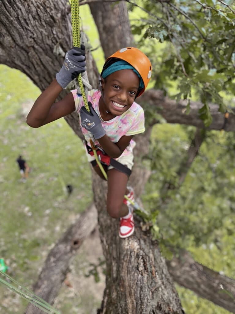 Smiling child wearing an orange helmet and safety harness climbs a large tree, holding onto a rope with gloves on.