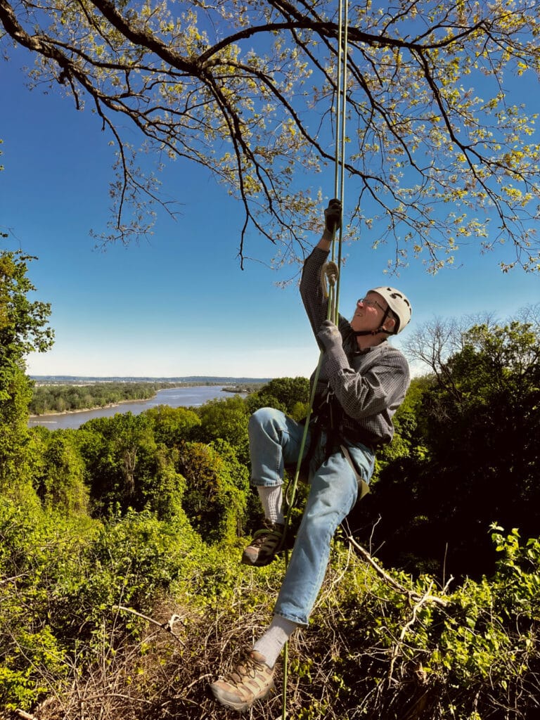 A person in a helmet and gloves climbing a rope system near the edge of a bluff, with a wide river and forest in the background.