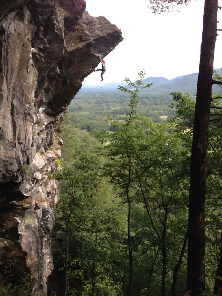 A climber hanging on an overhanging rock face high above a forest valley with mountains in the distance.
