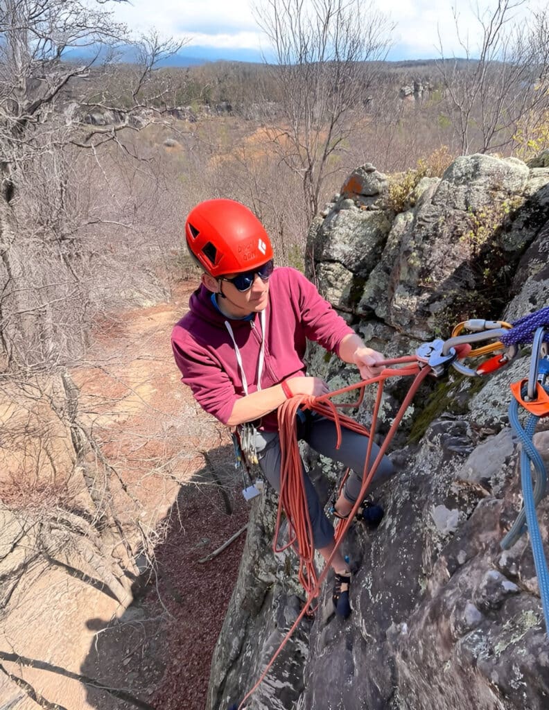 A climber in a red helmet and maroon hoodie setting up ropes on a rocky ledge with a forested valley below.