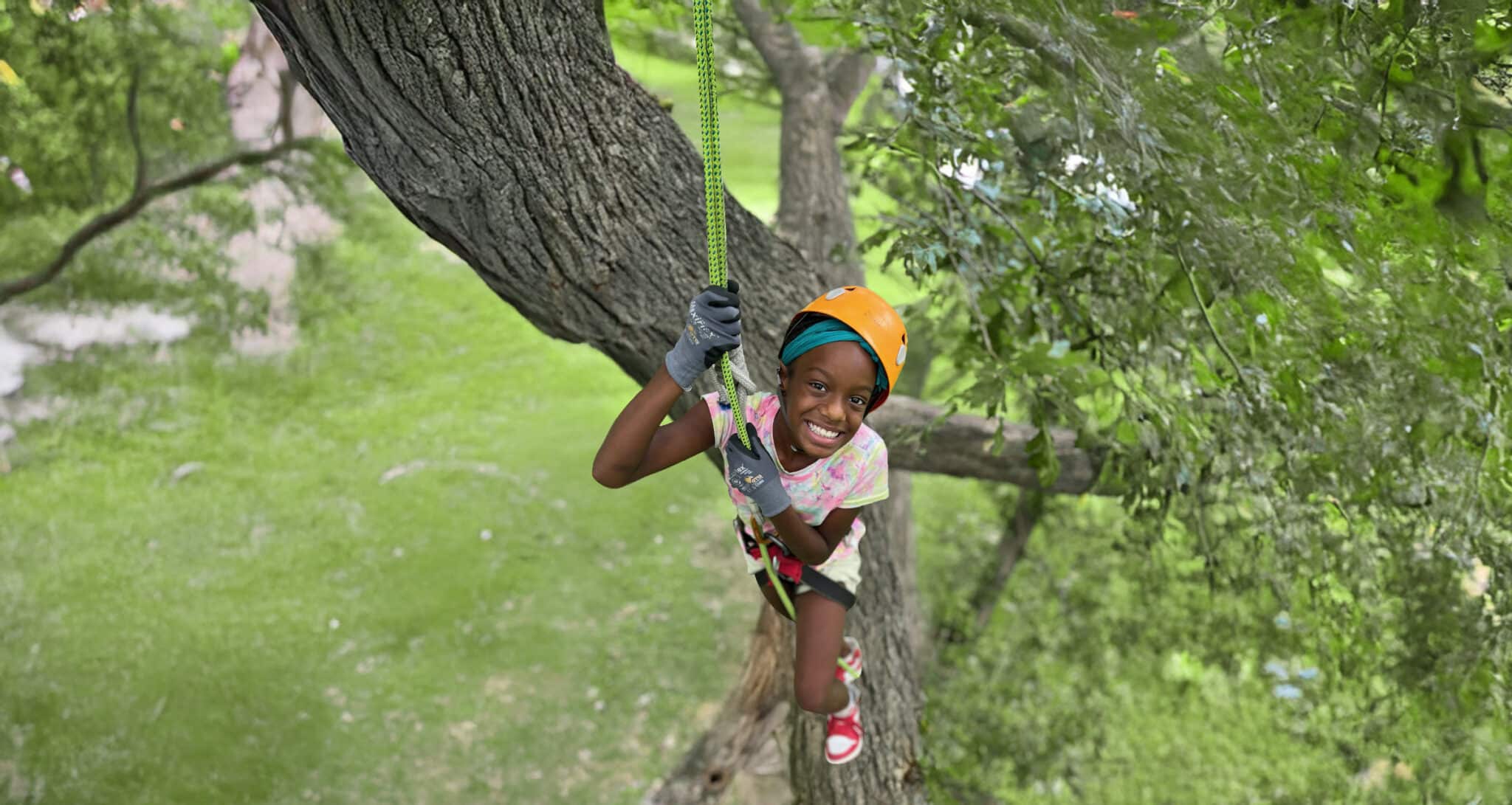 A smiling child in an orange helmet and gloves hanging from a rope in a large tree.