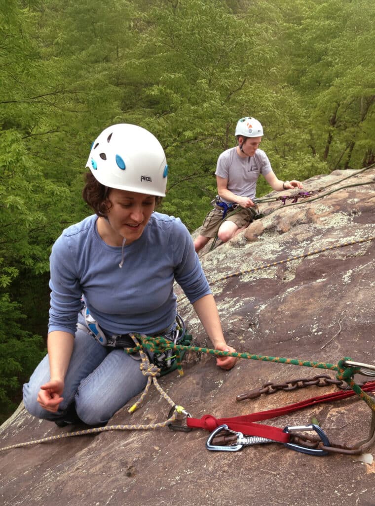 Two climbers wearing helmets and harnesses are positioned on a rocky ledge in a forested area. The climber in the foreground is kneeling while managing ropes connected to a multi-point anchor system built with slings, chains, and carabiners. A second climber sits slightly higher on the rock, also handling ropes. Dense green trees fill the background.