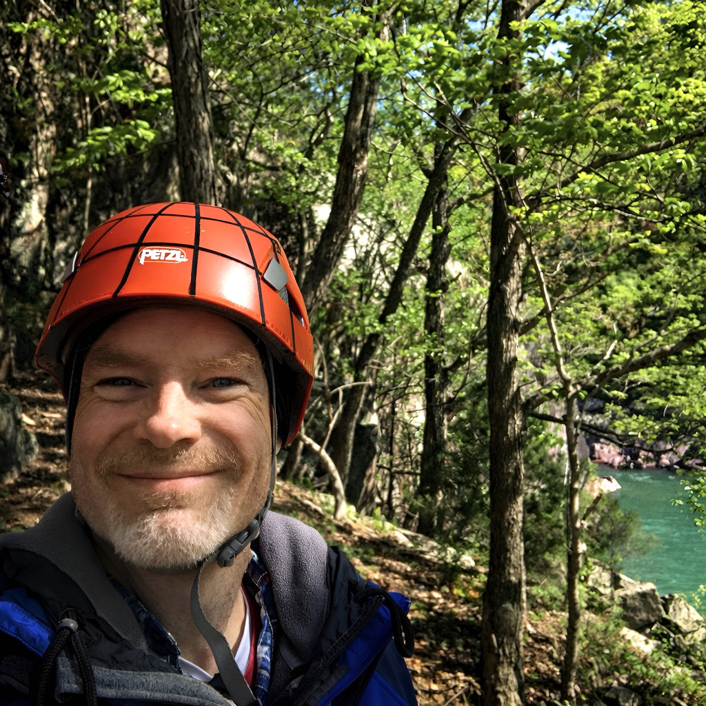 Smiling man wearing a red climbing helmet stands in a forested area with trees and a turquoise river visible in the background.
