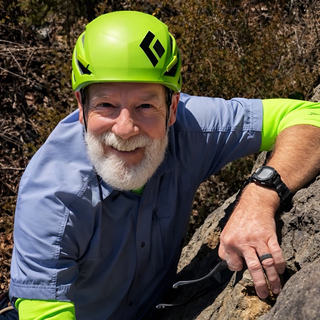 Smiling rock climber wearing a bright green helmet and blue shirt pauses on a rocky ledge, holding climbing gear in a wooded outdoor setting.