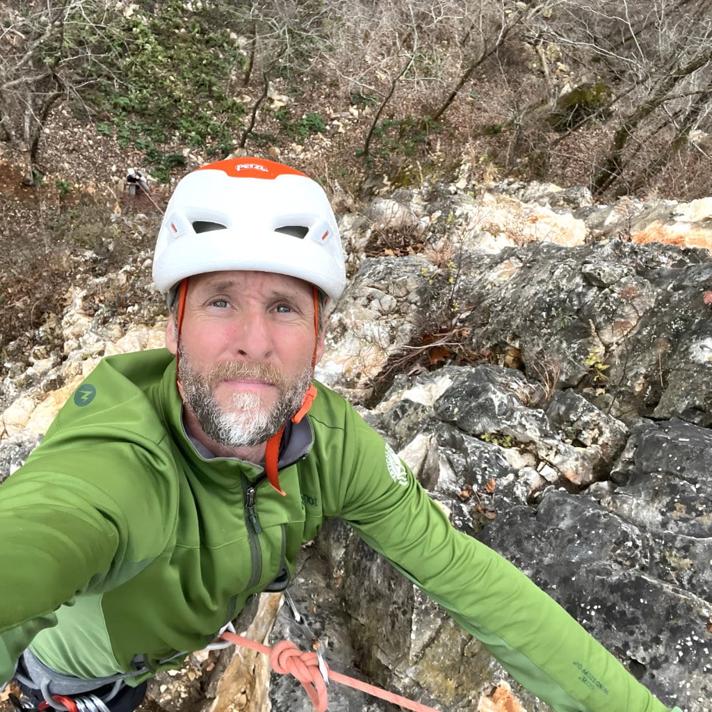 Rock climber wearing a white helmet and green jacket looks up from a steep rock face, secured by rope, with trees and exposed stone below.