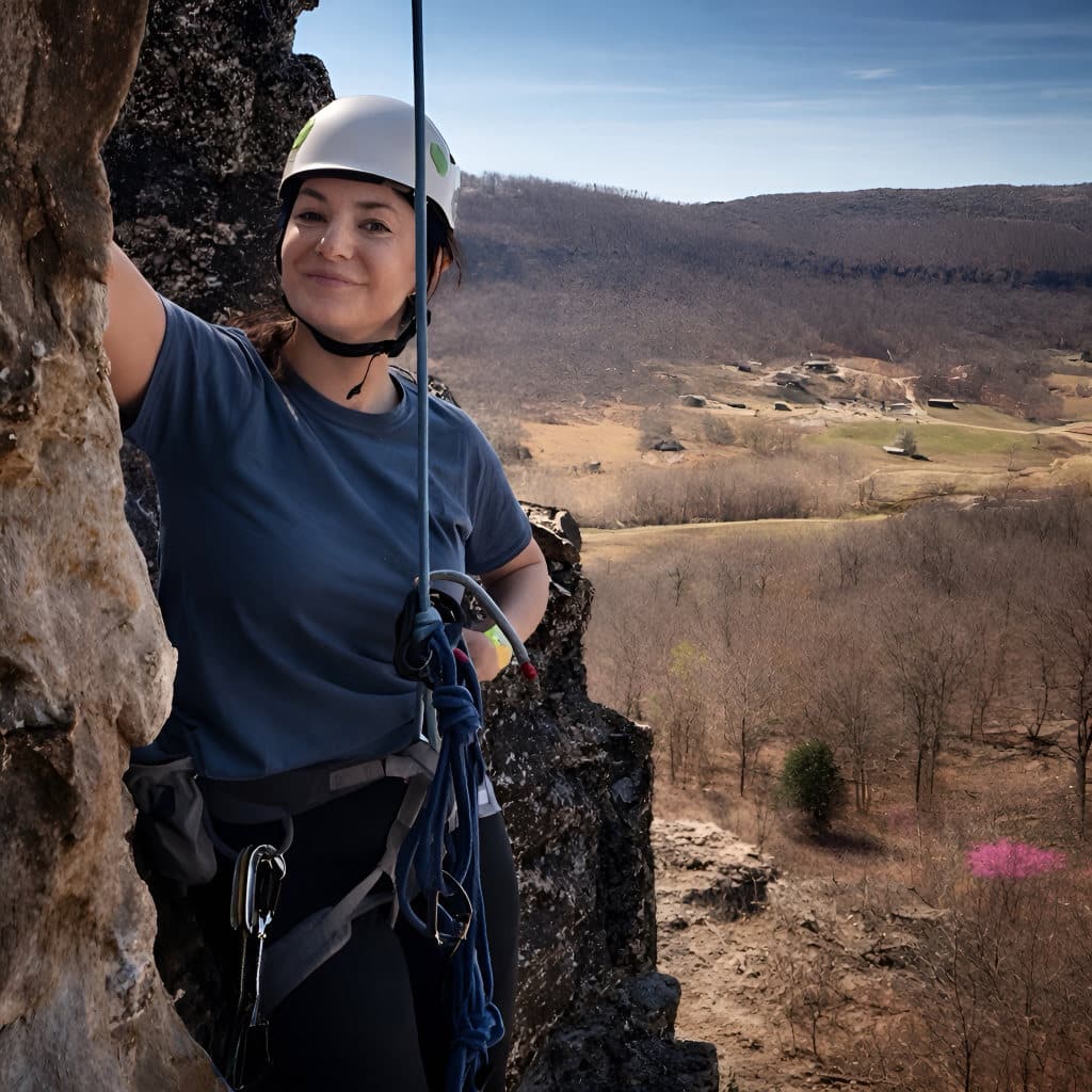 Smiling rock climber wearing a helmet and harness pauses on a cliff face, secured by rope, with a broad valley and hills visible in the background.