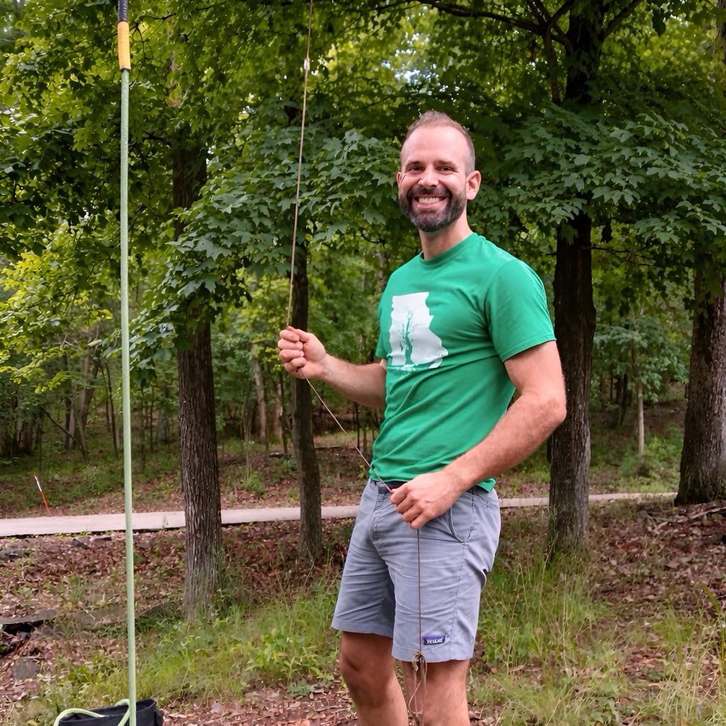 Smiling man wearing a green T-shirt and shorts stands in a wooded area holding a rope, with trees and a path visible behind him.