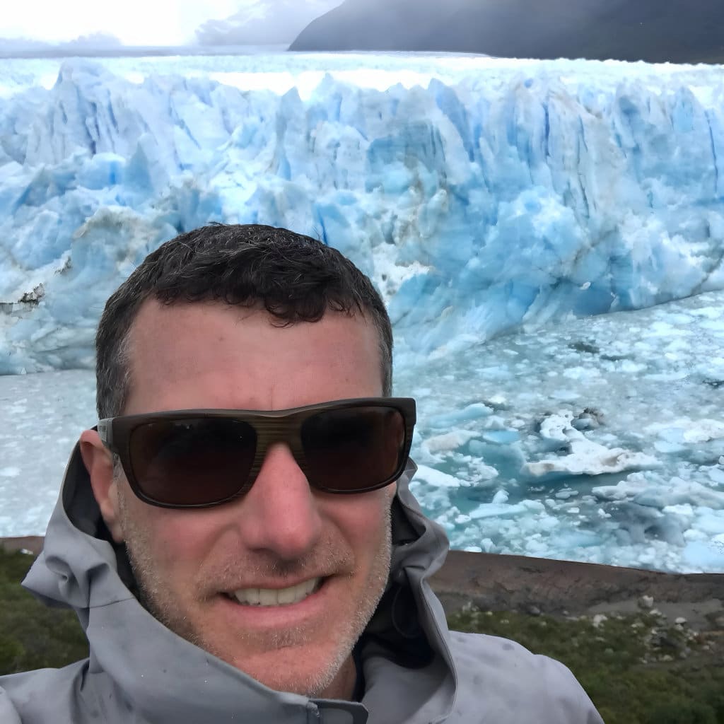 Man wearing sunglasses and a jacket stands in front of a massive blue glacier with ice and water visible below.
