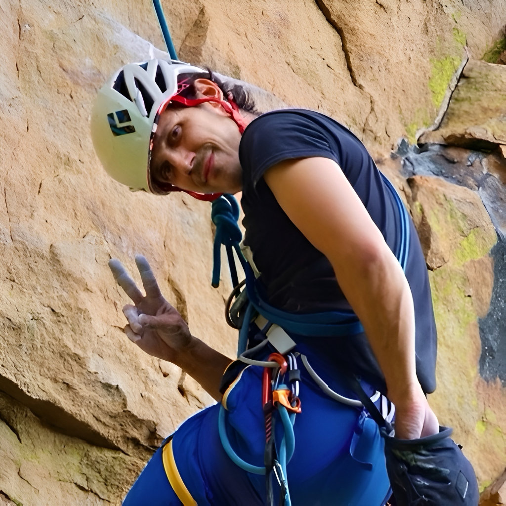 Smiling rock climber wearing a helmet, harness, and chalked gloves pauses on a rock face and flashes a peace sign while secured by a rope.
