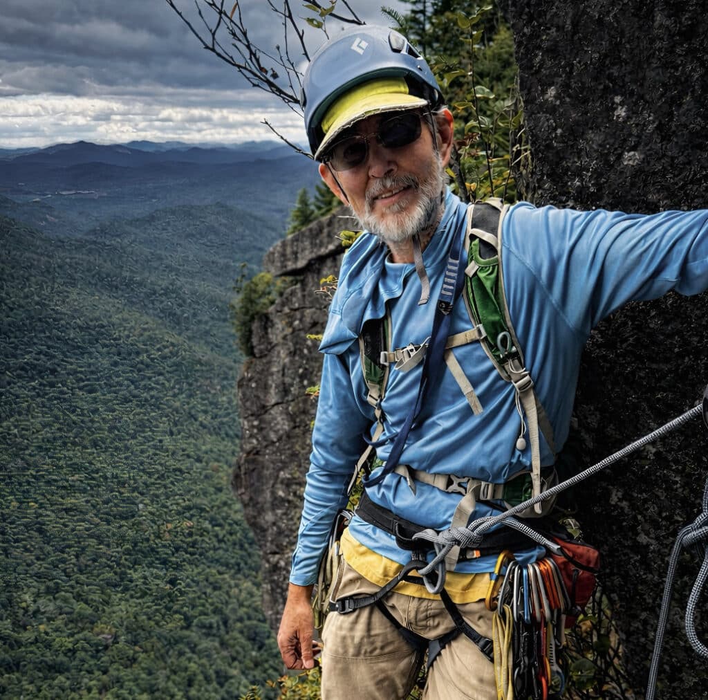 Older climber wearing a helmet, sunglasses, and safety harness pauses on a steep rock face overlooking a vast, forested mountain landscape under a cloudy sky.