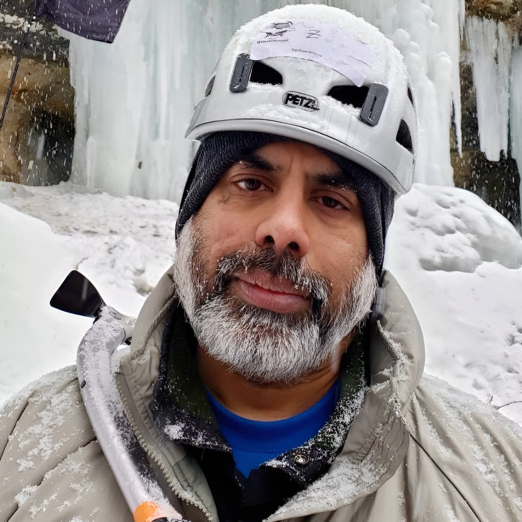 Bearded man wearing a climbing helmet and winter jacket stands in falling snow near frozen ice formations.